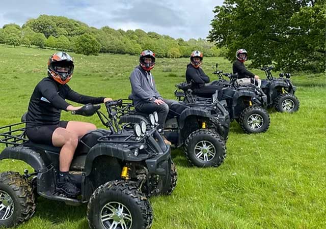 Four riders in helmets lined up on electric quad bikes in a grassy field, with trees behind, during a Cotswold activity experience day tour