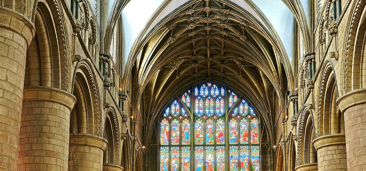 Massive Norman columns lining the nave of Gloucester Cathedral, leading to the vaulted ceiling and great stained glass window